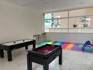 Une pièce avec deux tables de baby-foot, un tapis de jeu coloré et une fenêtre donnant sur des immeubles.