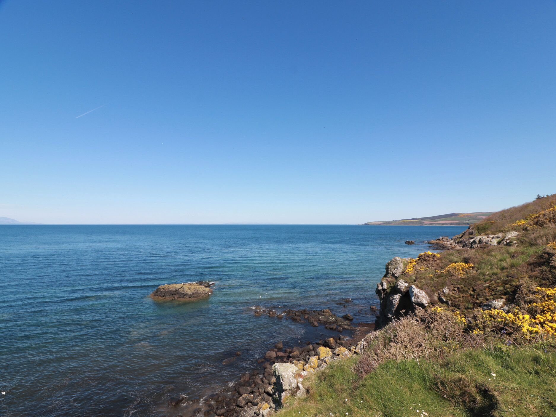 A coastal landscape with clear skies, calm sea, rocky shoreline, and sparse vegetation.