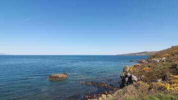 A coastal landscape with clear skies, calm sea, rocky shoreline, and sparse vegetation.