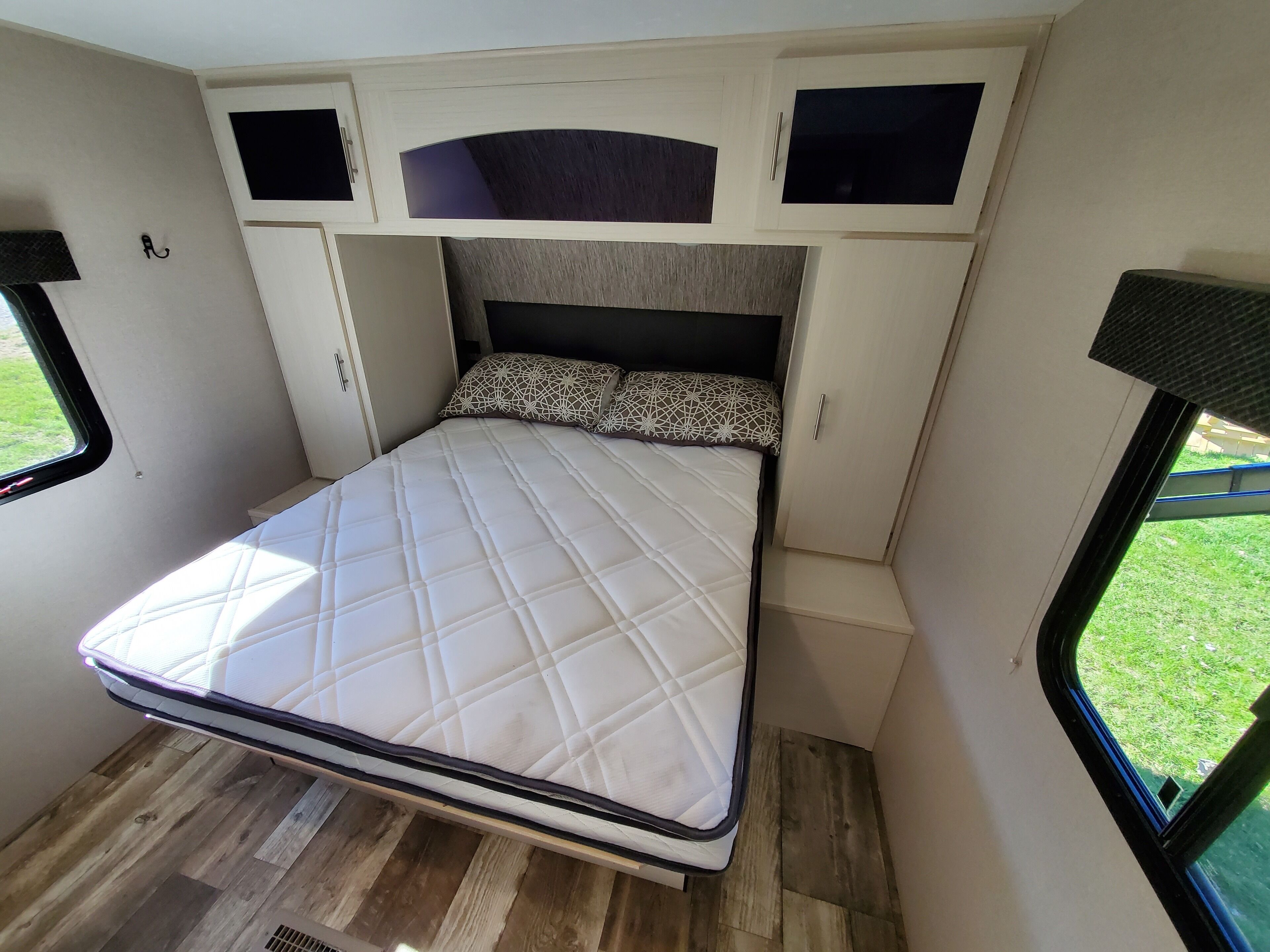 A compact bed with a quilted mattress, a headboard with a dark panel, and a window with a view of greenery.