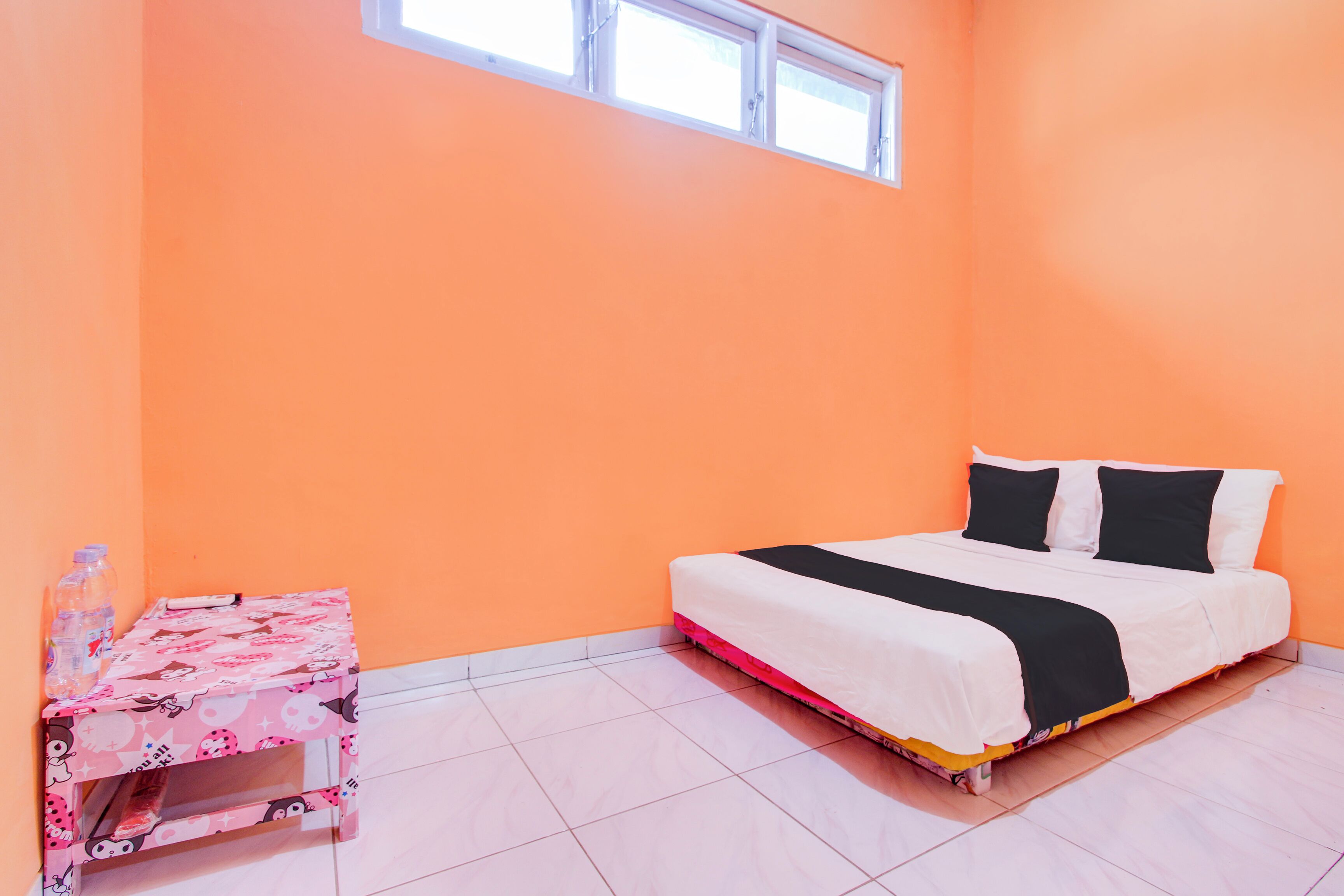 A room with an orange wall, a bed with black and white bedding, a pink table with cartoon patterns, and a window above the bed.