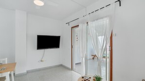 Een moderne woonkamer met een flatscreen televisie aan de muur, een eethoek met tafel en stoelen, en een schuifdeur met transparante gordijnen.
