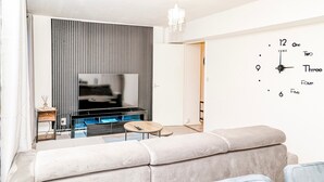A modern living room with a sofa, a TV, and a wall clock. - Kingston Duplex Apt Balcony, Parking & Wi-fi (Kingston Upon Thames)