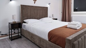 A neatly made bed with white bedding and a brown throw, a bedside table with a lamp, and a window with curtains. - Kingston Duplex Apt Balcony, Parking & Wi-fi (Kingston Upon Thames)