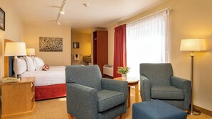 Ett hotellrum med en säng, två fåtöljer, ett sidobord, en lampa, ett litet bord med en växt och ett fönster med gardiner.