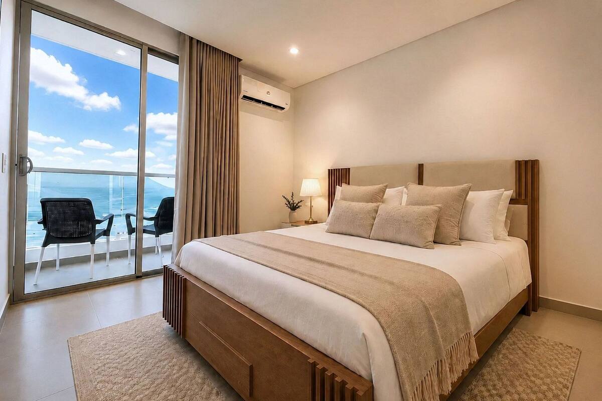 A modern hotel room with a large bed, a balcony with chairs, and a view of the sea.