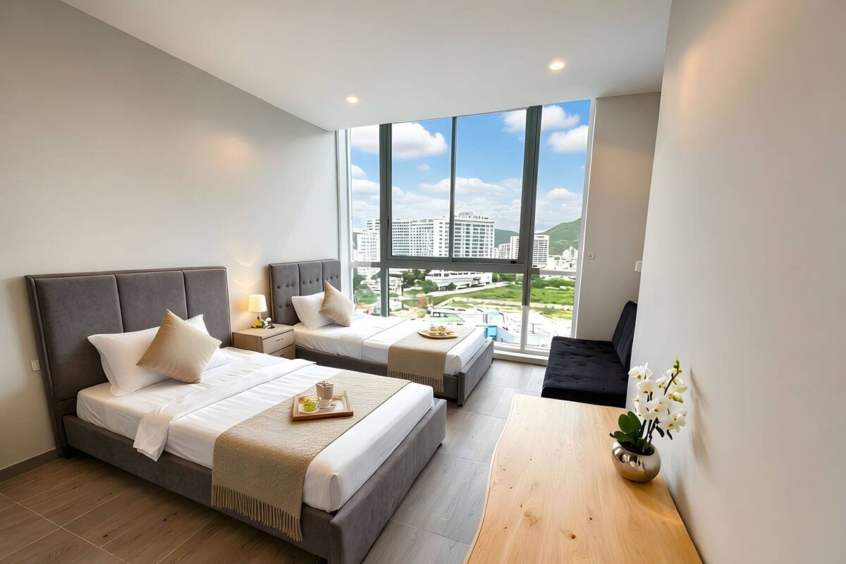 A modern hotel room with two beds, a small table, and a view of the cityscape.