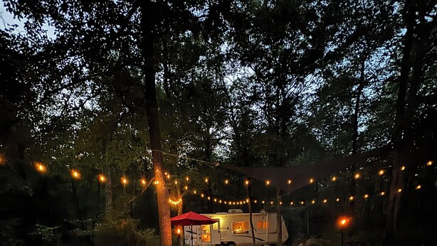 Un espace extérieur aménagé avec des chaises rouges et un foyer, entouré de rochers et d’arbres, le tout illuminé par des guirlandes lumineuses suspendues au-dessus.