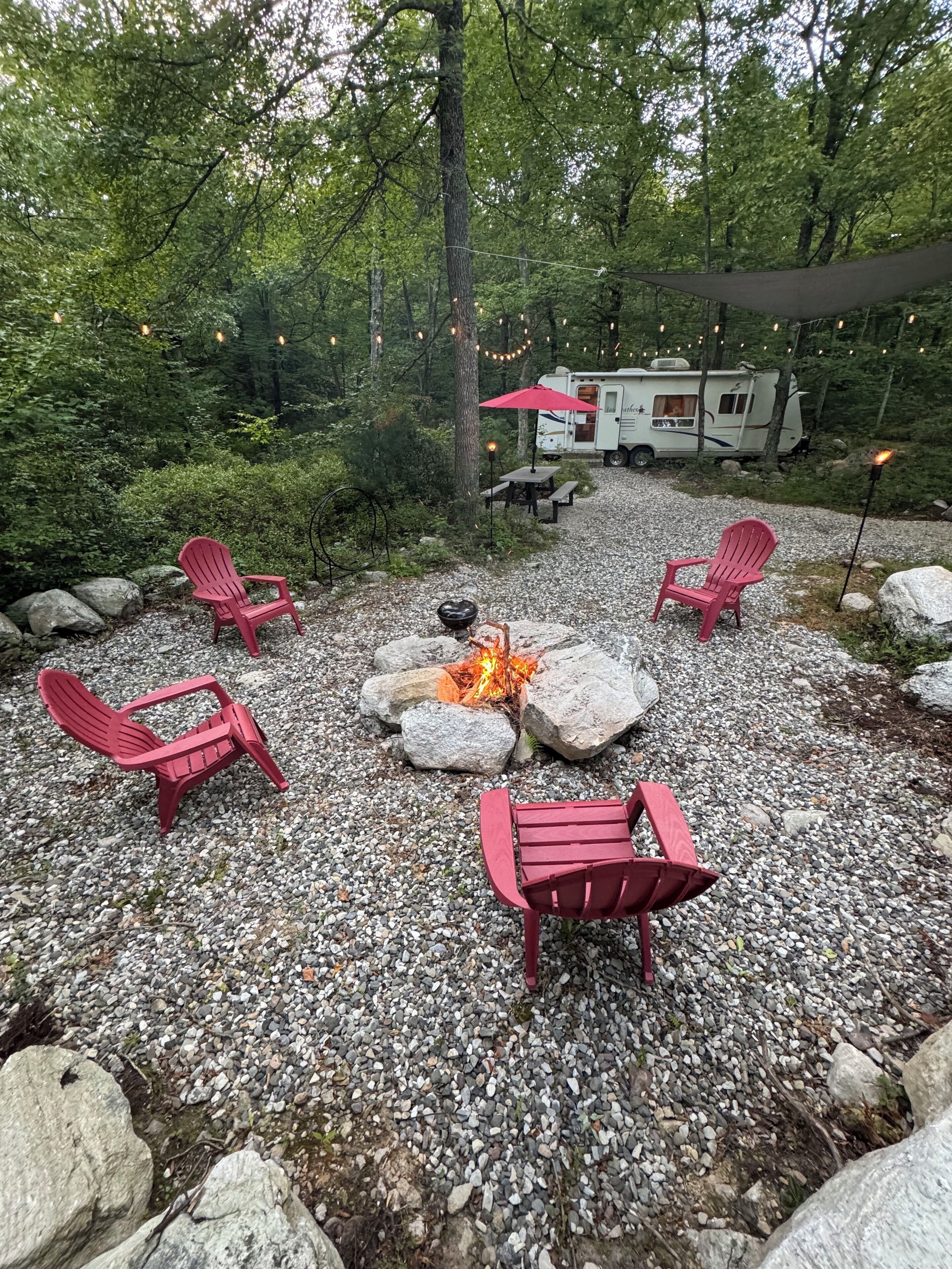 Een camping met een vuurkuil, omringd door rode Adirondack stoelen, een eettafel en een rode parasol.