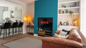 Una sala moderna con una pared de acento color turquesa, un televisor de pantalla plana, un sofá de cuero marrón, una zona de comedor con sillas negras y una cocina al fondo.