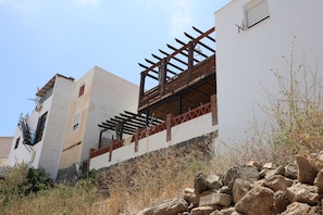 A building with a white facade, a wooden pergola on the upper floor, and a stone wall in the foreground.