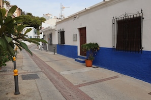A narrow street with buildings painted in white and blue, a potted plant, and a metal bollard.