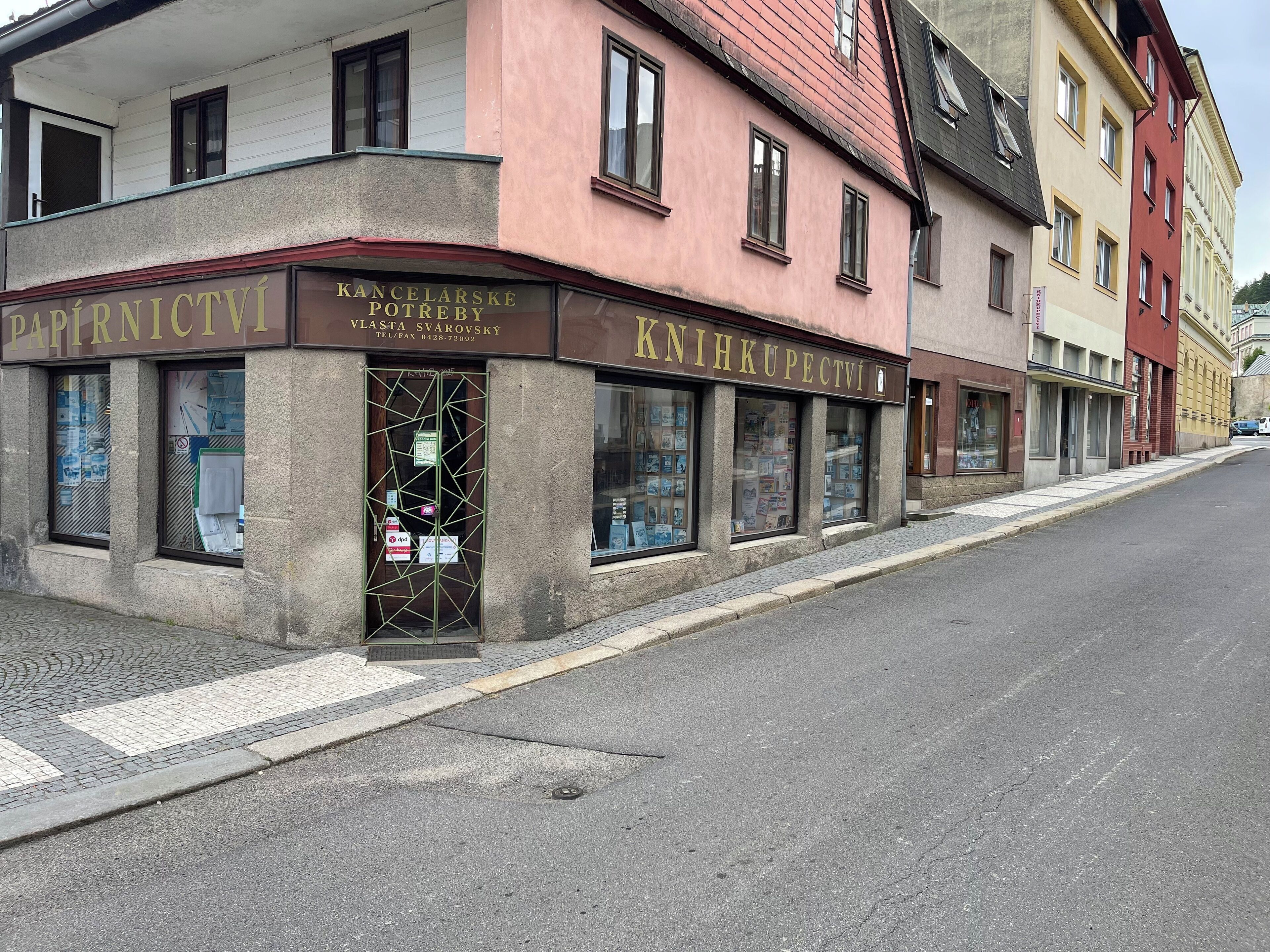 Une vue d’une rue, montrant un immeuble avec deux devantures de magasins, l’une portant l’inscription « PAPIRNICTVI » et l’autre « KNIHKUPECTVI ».