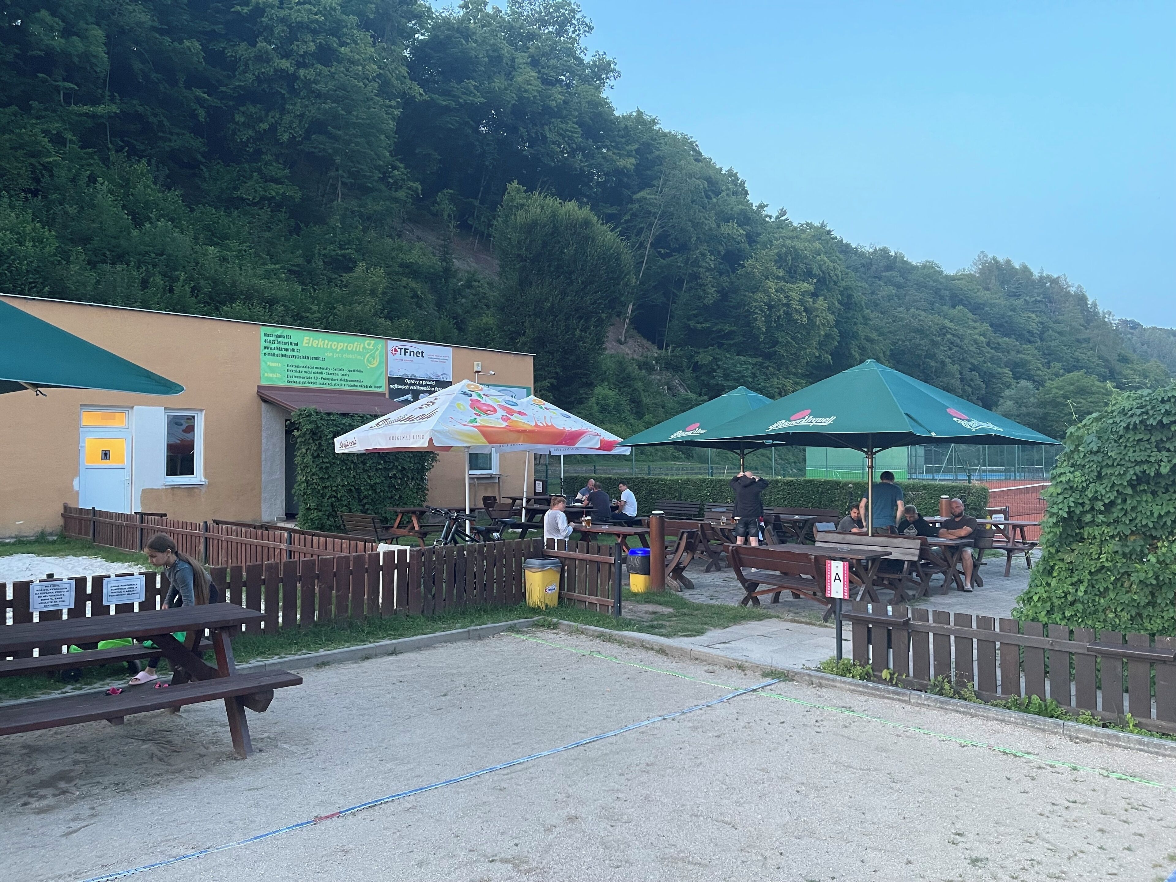 Ein Außenbereich mit Picknicktischen und Sonnenschirmen, umgeben von einem Holzzaun, mit einem Gebäude im Hintergrund.