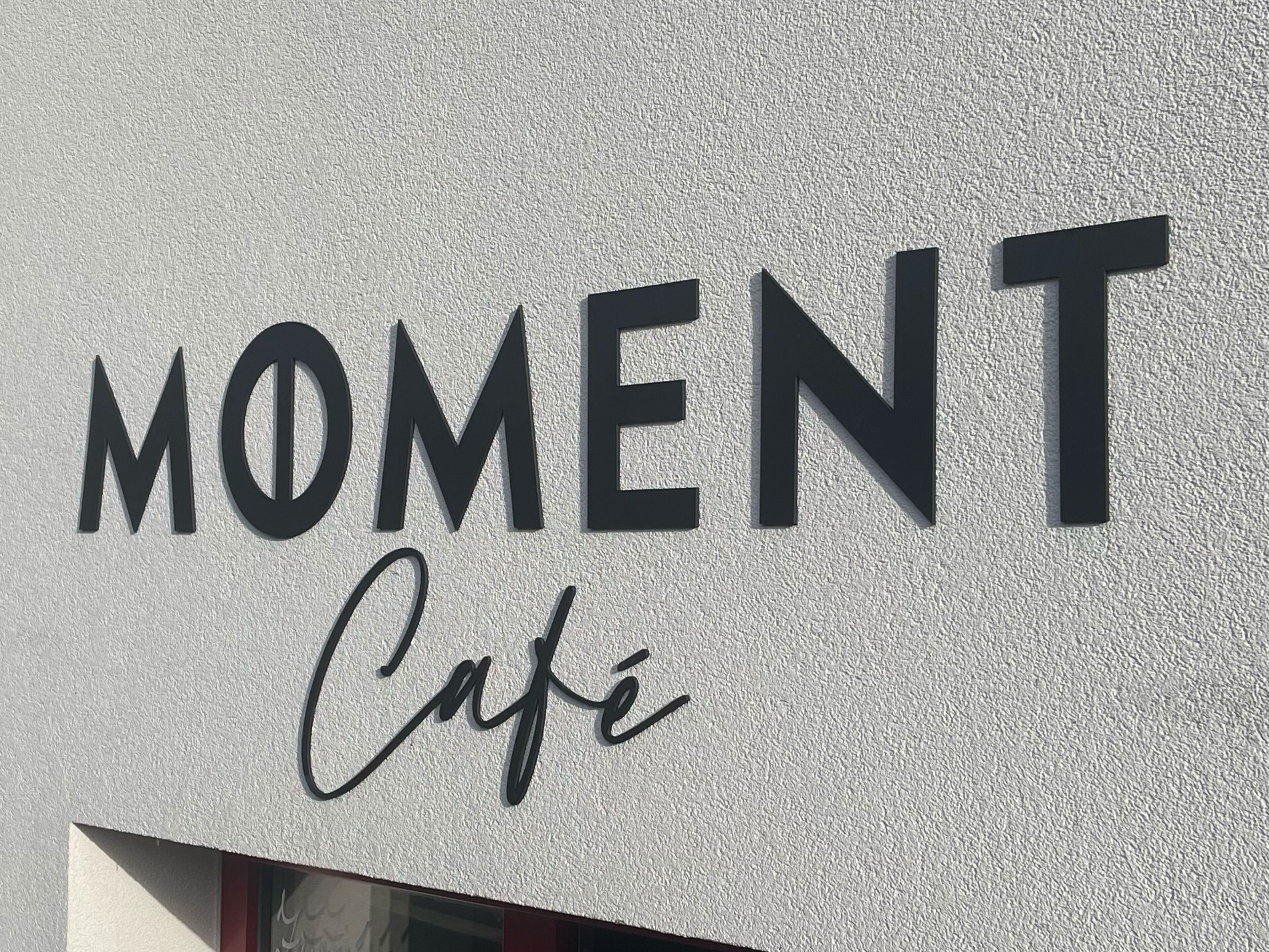 Ein Schild mit den Aufschriften „MOMENT“ und „Cafe“ an einer strukturierten Wand.