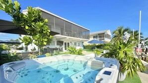 Un jacuzzi con zona de estar integrada, rodeado de vegetación exuberante y un edificio moderno al fondo.