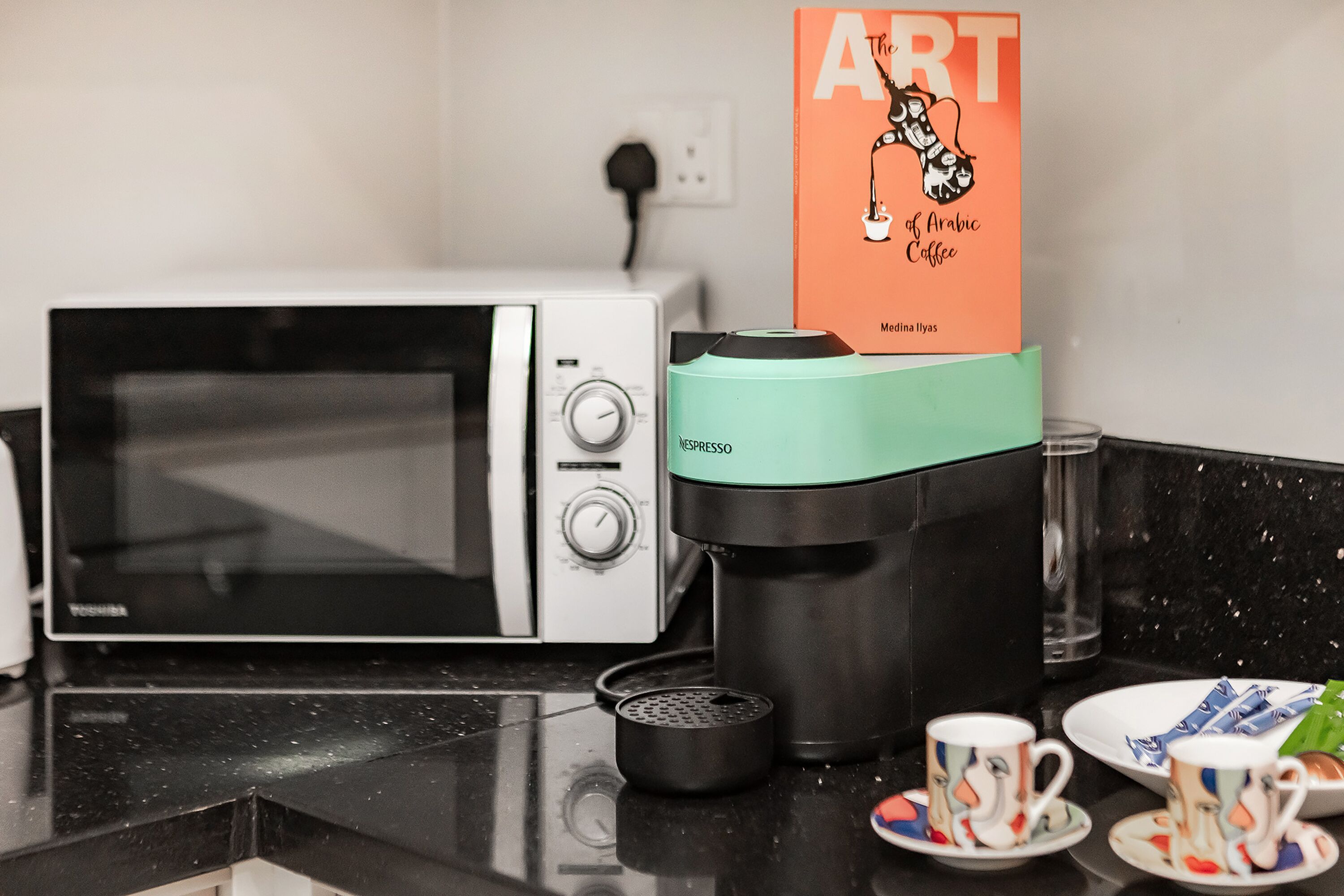 Eine Küche mit einer Mikrowelle, einer Kaffeemaschine und einem Buch mit dem Titel 'The Art of Arabic Coffee' von Medhina Ilyas.