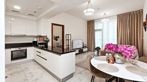 A modern kitchen with white cabinets, a black countertop, and a built-in oven. There is a dining area with a white table and chairs, and a vase of pink flowers. - Stylish 1BR Apartment at Meera Tower (Dubai)