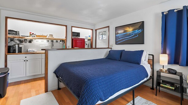 A compact hotel room with a bed, a small bedside table with a lamp, a view of the kitchen, and a framed picture on the wall.