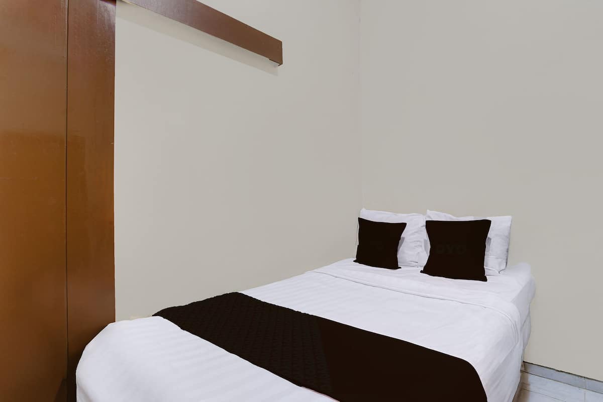 A single bed with white bedding and two black pillows, a wooden wardrobe, and a plain white wall.