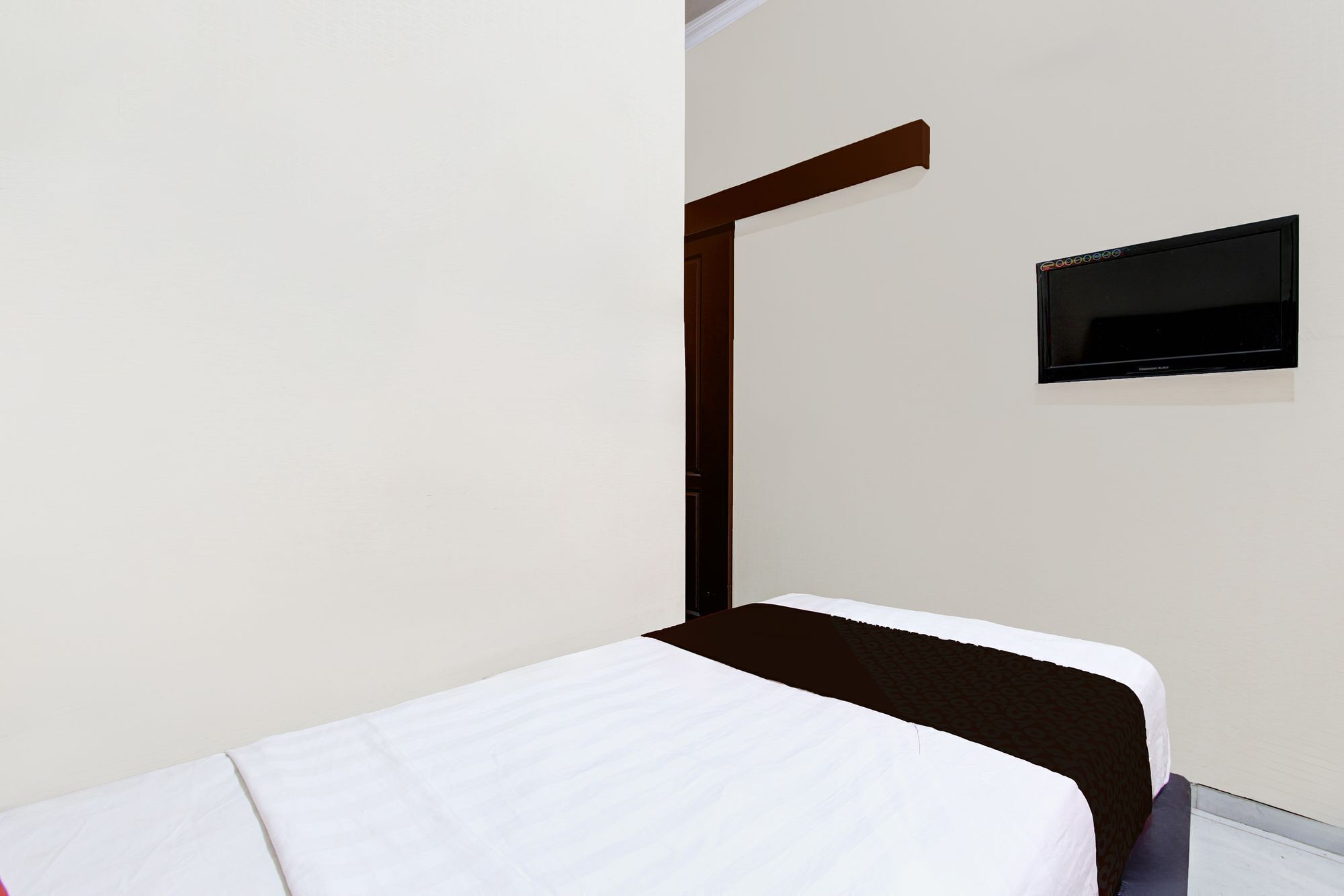 Un lit simple avec un matelas blanc et une tête de lit marron, dans une pièce dont le mur est blanc et où est fixé un petit téléviseur noir.