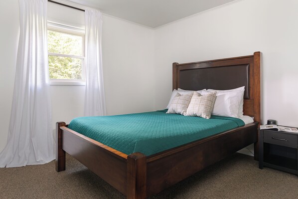 Una cama bien hecha con estructura de madera y cubrecama verde, una mesita de noche con lámpara, y una ventana con cortinas blancas.