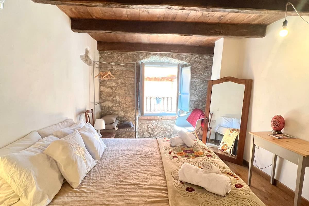 A cozy bedroom with a wooden ceiling, a stone wall, a bed with floral bedding, a bedside table with a lamp, and a mirror.
