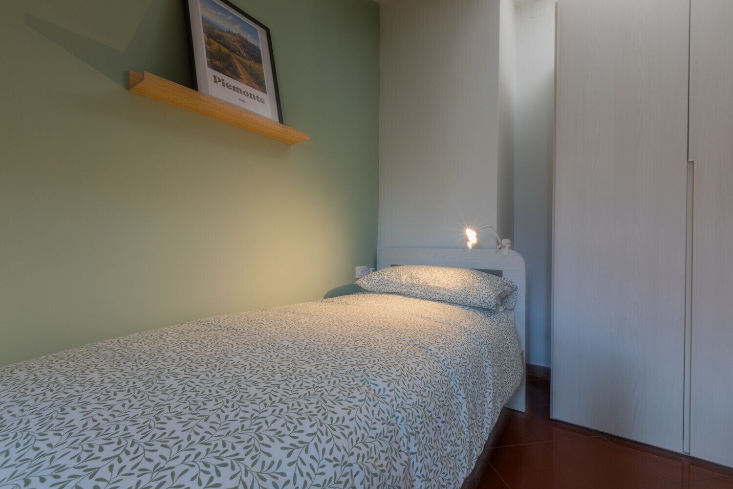 Una cama pequeña y bien hecha con una colcha estampada, un cuadro enmarcado en un estante arriba y un armario blanco a la derecha.
