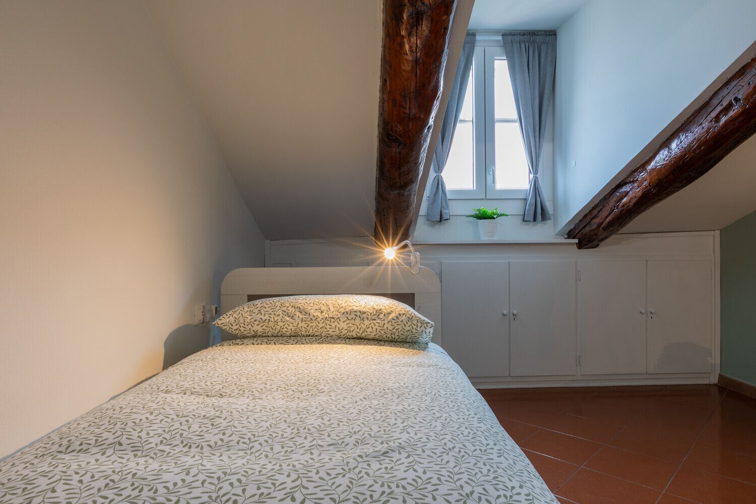 A small, cozy bedroom with a sloped ceiling, a bed with patterned bedding, a window with curtains, and a wooden beam.