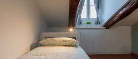 A small, cozy bedroom with a sloped ceiling, a bed with patterned bedding, a window with curtains, and a wooden beam.