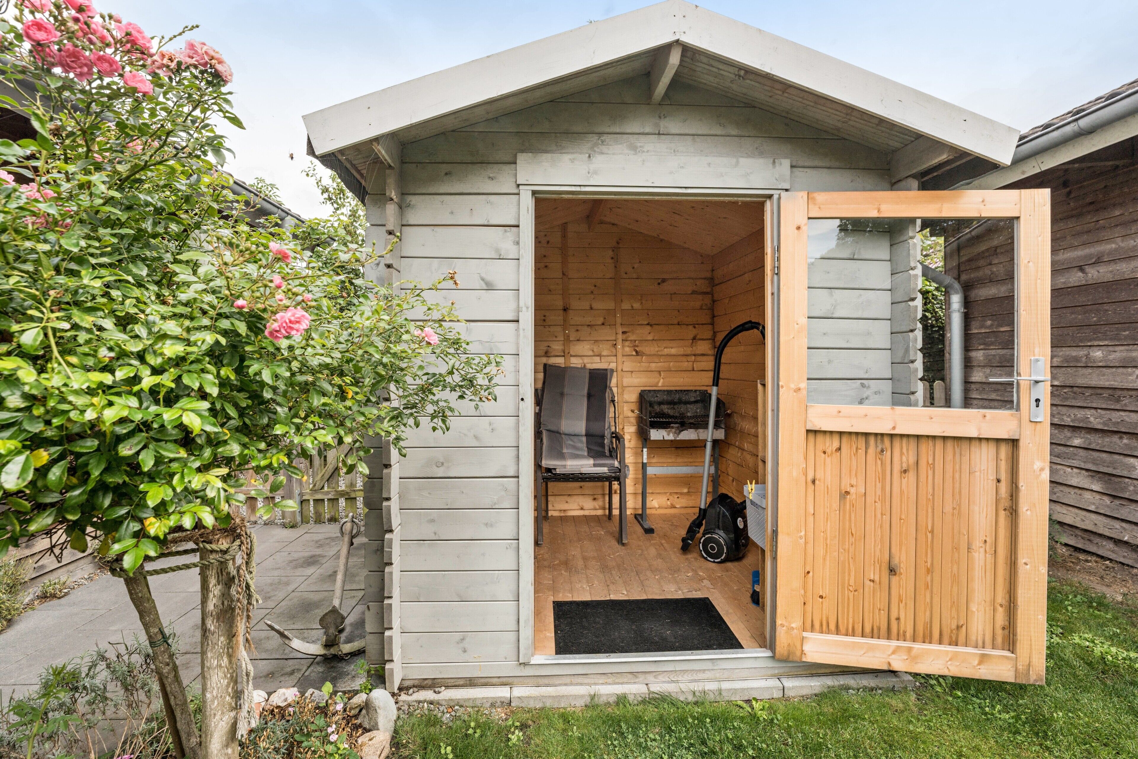 Una cabaña de madera con la puerta abierta, mostrando una zona de estar con una silla, una mesa, una parrilla negra y una cerca de madera.