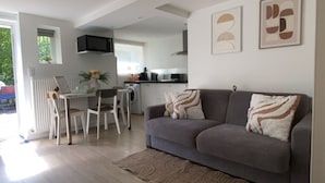 A modern living room with a grey sofa, two throw pillows, a dining table with chairs, a kitchen area in the background, and a large window providing natural light. - New / Oasis in the Heart of Nancy with Garden and Free Parking 🍃 (Nancy)