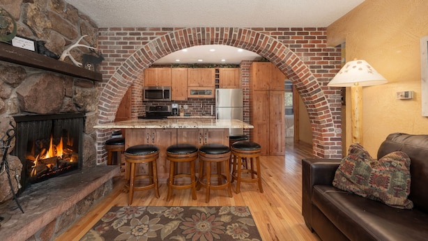 A kitchen with a stone fireplace, wooden bar stools, and a brick archway leading to the dining area.