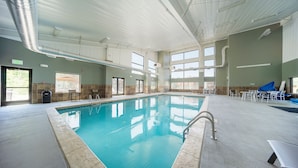 An indoor swimming pool with clear water, surrounded by a tiled floor and walls. There are windows allowing natural light, and a few chairs and tables are visible.
