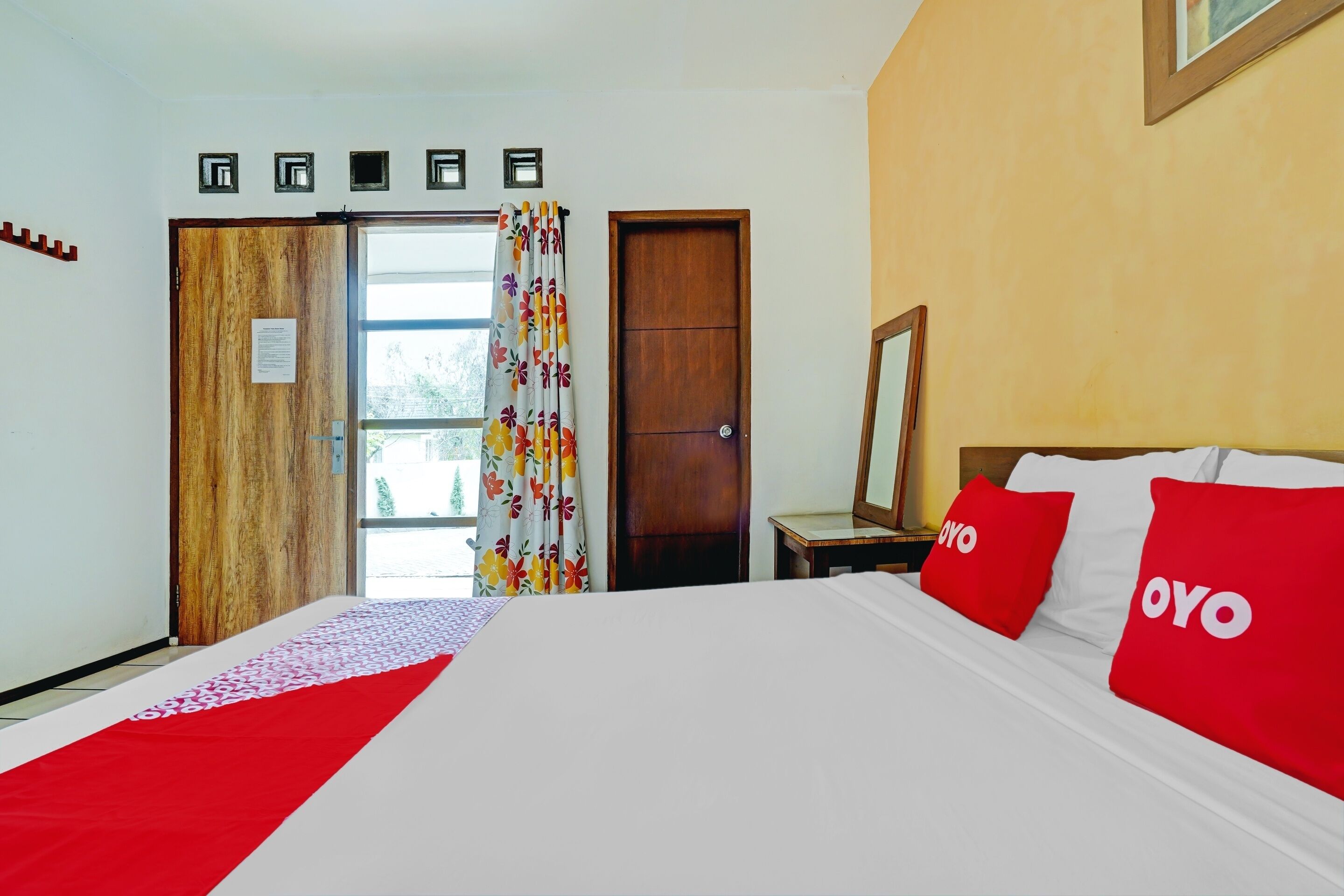 A hotel room with a large bed, a wooden door, a window with curtains, and a nightstand with a mirror.