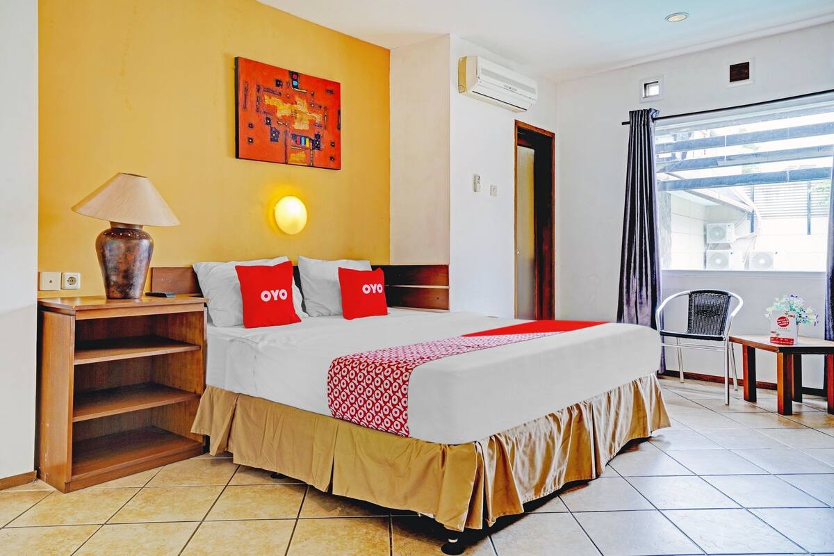 Une chambre d’hôtel comprenant un lit, une table de chevet, une lampe, un climatiseur fixé au mur, une fenêtre avec des rideaux et une petite table sur laquelle est posé un vase.
