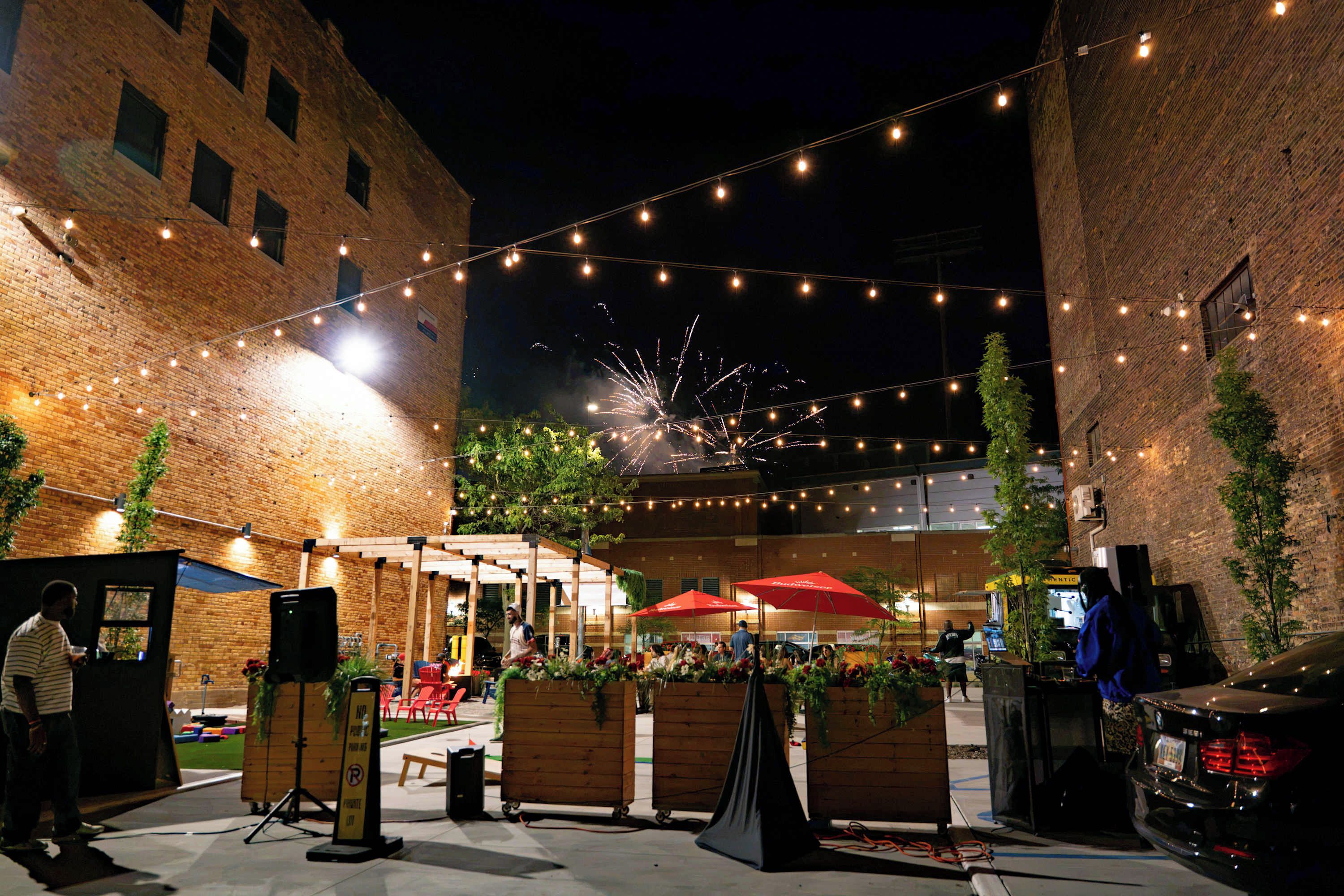 An outdoor seating area with string lights, wooden planters, and a brick building in the background.