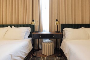 A hotel room with two beds, a desk, two lamps, and a striped ottoman.