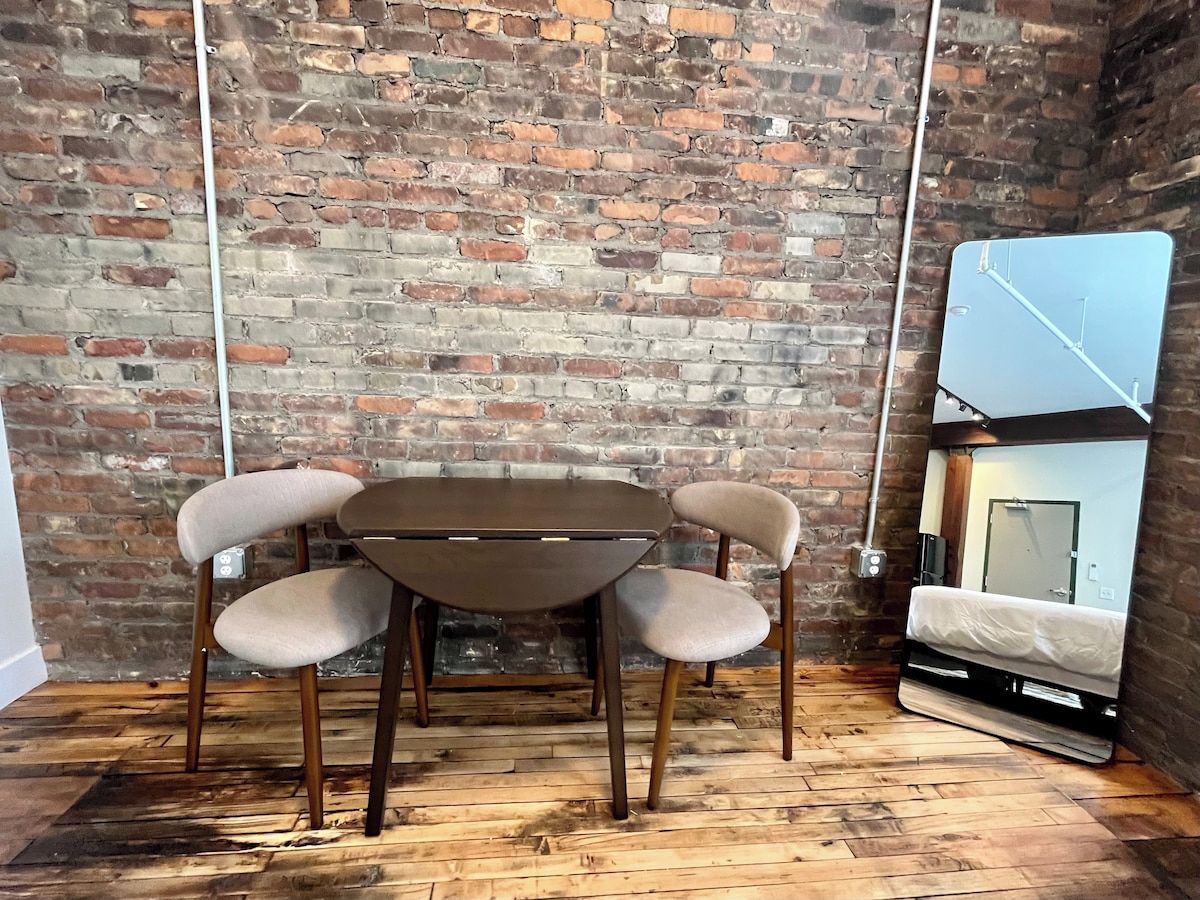 Une pièce avec un mur en briques, un plancher en bois, une table ronde avec quatre chaises et un lit avec un matelas et des oreillers.