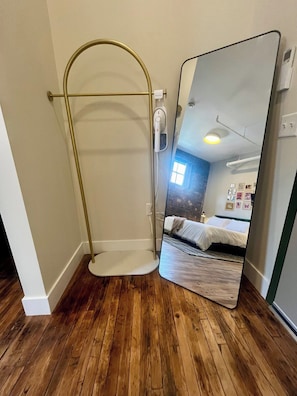 Una habitación con suelo de madera, ducha con mampara dorada, inodoro blanco y un espejo grande que refleja una cama y una ventana.