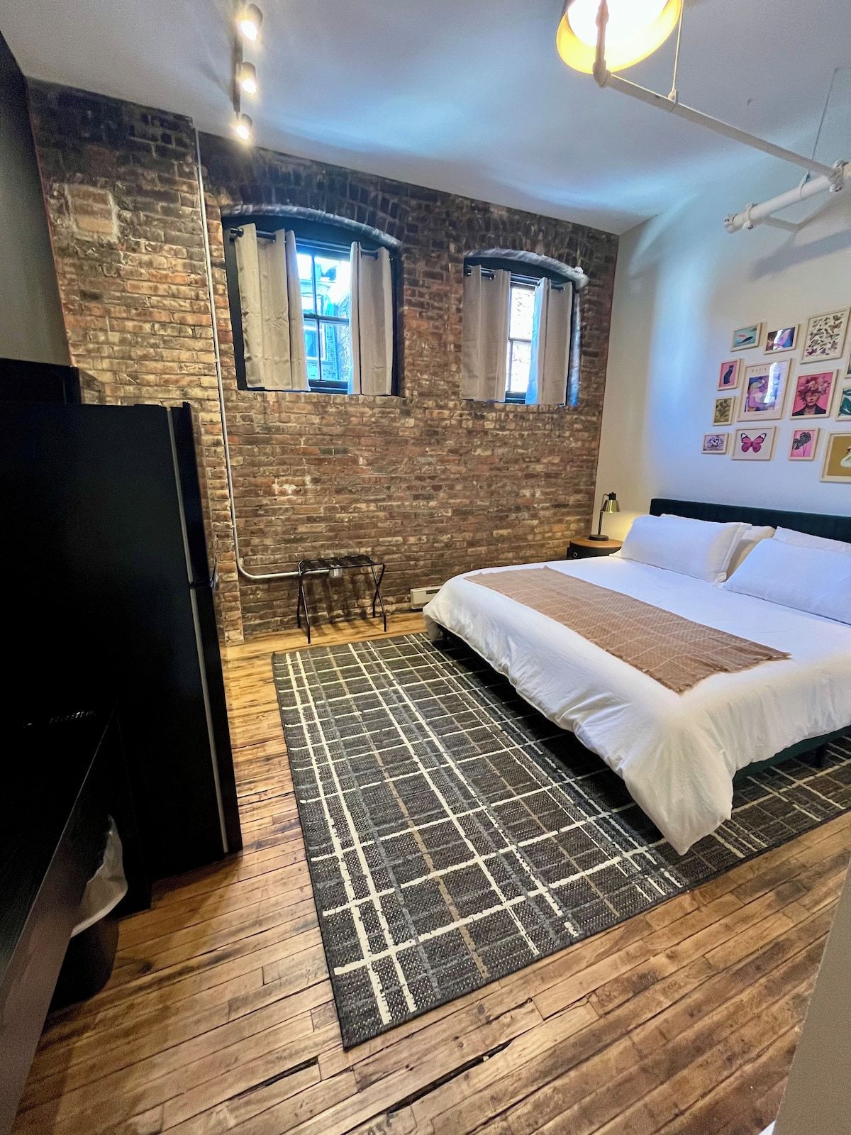 Ein Schlafzimmer mit Backsteinwand, einem Bett mit weißer Bettwäsche, einem schwarzen TV-Schrank und einem gemusterten Teppich.