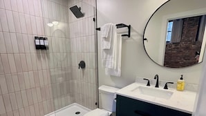 A modern bathroom with a glass shower enclosure, a toilet, a sink with a mirror, and dark-toned cabinets. - Toledo King Loft | Ballpark & Dining | Suite 203 (Toledo)