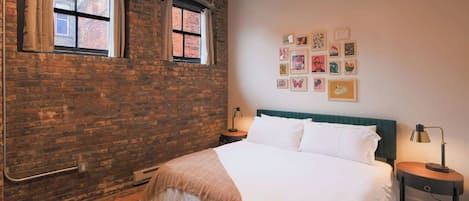 Un dormitorio con pared de ladrillo, una cama con ropa de cama blanca, un cabecero, una mesita de noche, una lámpara y una pared decorada con cuadros.