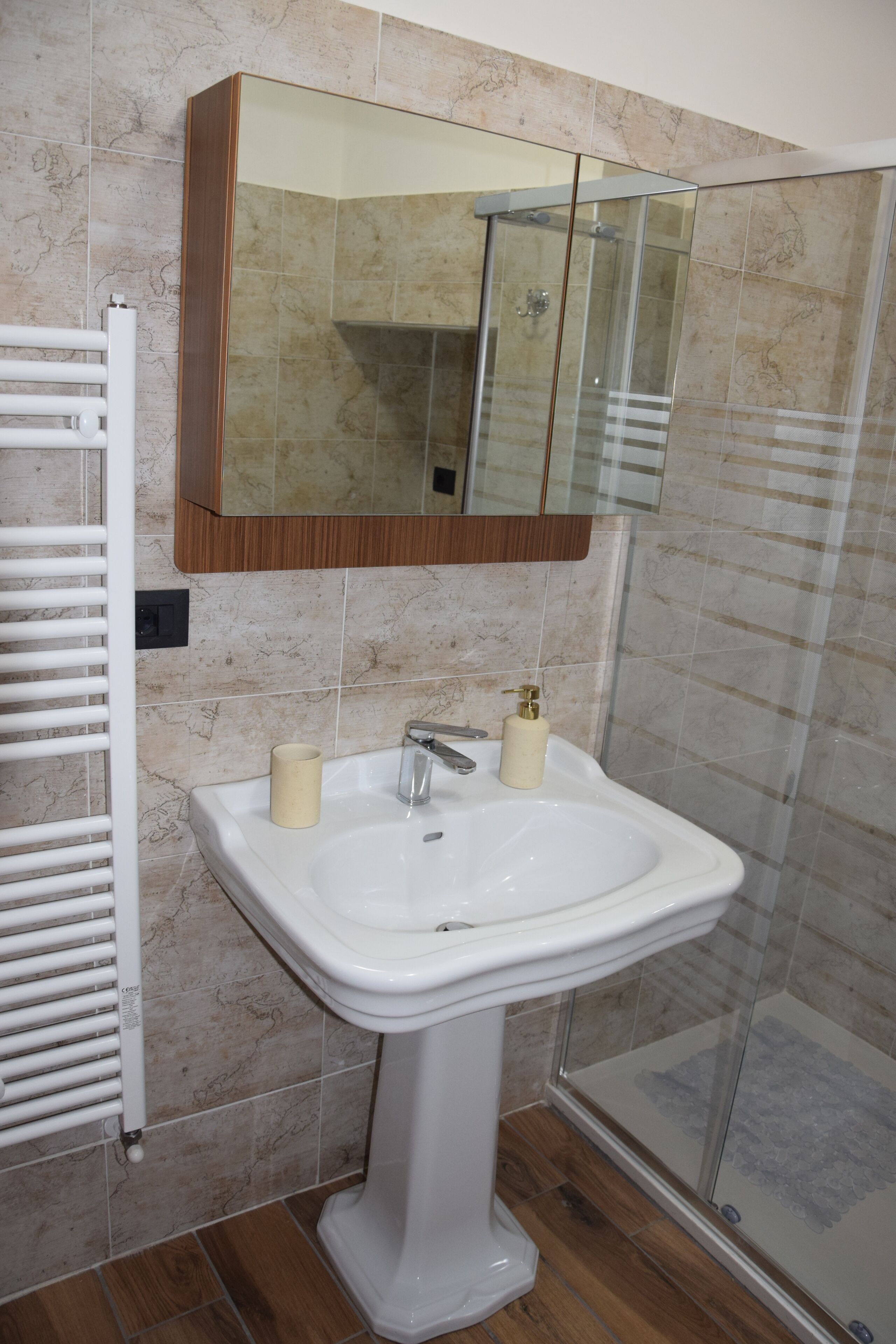 Un baño con un lavamanos blanco de pedestal, una ducha con cerramiento de vidrio y un espejo con marco de madera sobre el lavamanos.