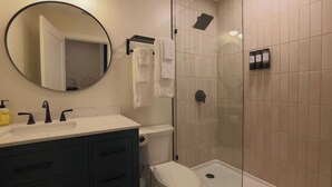 A bathroom with a round mirror, a sink with black faucets, a toilet, a shower with a glass door, and a towel rack with white towels. - Toledo Loft | Walk to Ballpark | Suite 204 (Toledo)