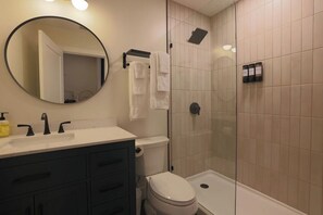 A bathroom with a round mirror, a sink with black faucets, a toilet, a shower with a glass door, and a towel rack with white towels. - Toledo Loft | Walk to Ballpark | Suite 204 (Toledo)