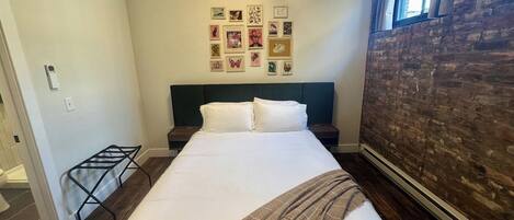 Un dormitorio con cama, cabecera, pared con cuadros, ventana y pared de ladrillo.