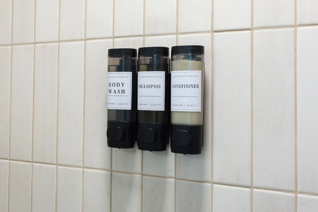Three toiletries dispensers labeled 'BODY WASH,' 'SHAMPOO,' and 'CONDITIONER' mounted on a tiled wall.