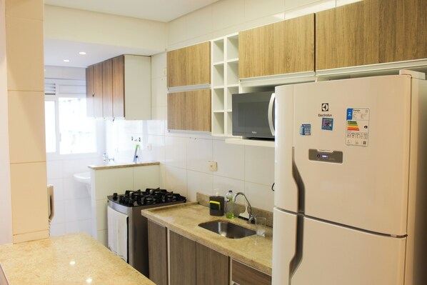 A modern kitchen with wooden cabinets, a built-in microwave, a refrigerator, a gas stove, and a sink. - 2 Min da Praia - 2Q - Vaga de Garagem (Santos)