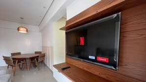 A modern living room with a wooden TV stand, a dining area with a round table and chairs, and a ceiling light fixture. - 2 Min da Praia - 2Q - Vaga de Garagem (Santos)